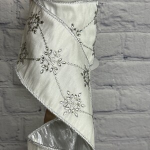 May include: A roll of white ribbon with silver snowflake embellishments. The ribbon has a satin finish and is trimmed with silver edging. The snowflakes are detailed with sparkling accents, perfect for holiday decorations or gift wrapping.