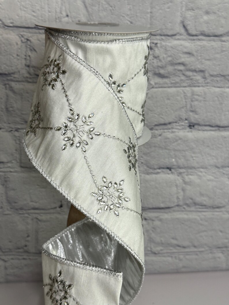 May include: A roll of white ribbon with silver snowflake embellishments. The ribbon has a satin finish and is trimmed with silver edging. The snowflakes are detailed with sparkling accents, perfect for holiday decorations or gift wrapping.