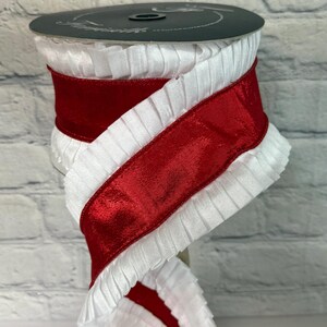 May include: A spool of white pleated ribbon with a wide, shiny red satin ribbon wrapped around it. The ribbon is displayed against a white brick background. The spool is dark gray.