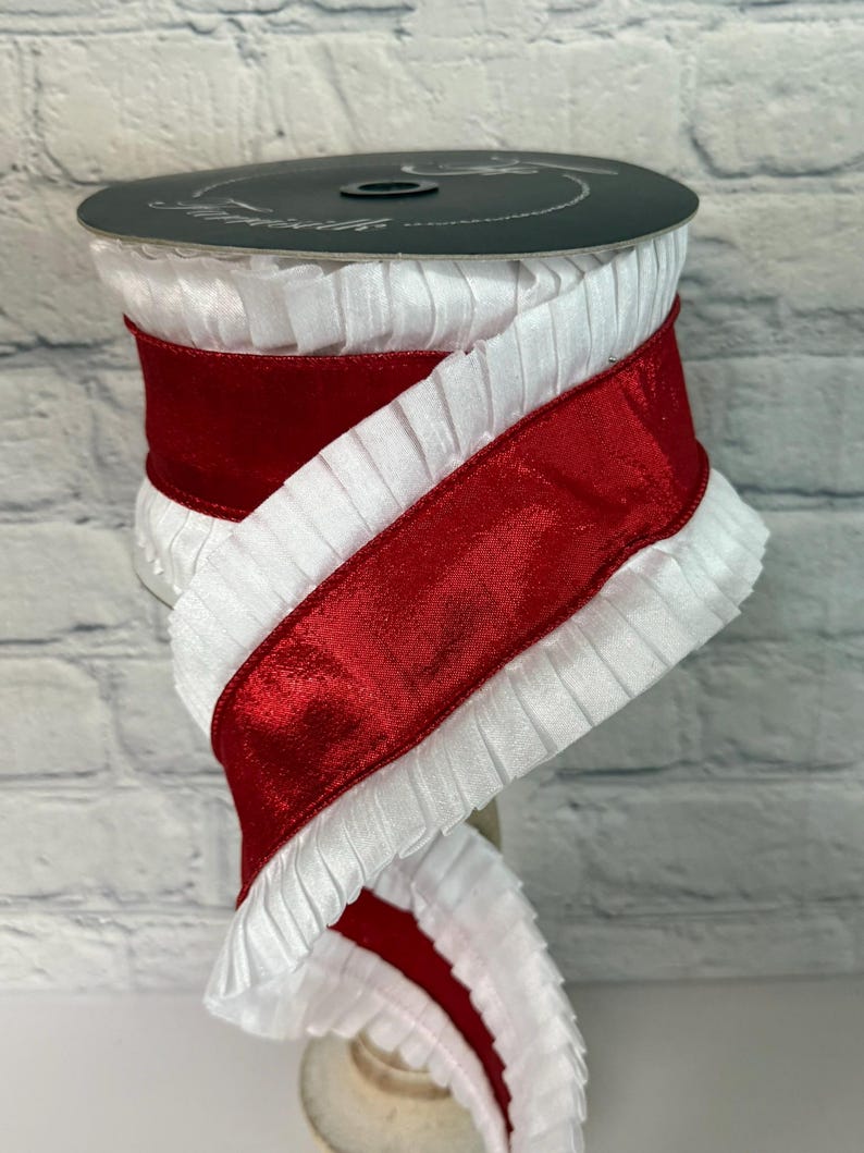 May include: A spool of white pleated ribbon with a wide, shiny red satin ribbon wrapped around it. The ribbon is displayed against a white brick background. The spool is dark gray.