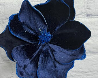 May include: A large, navy blue velvet flower with glitter edging and a textured center. The flower has multiple petals and is displayed in a clear glass vase. The flower is a decorative item.