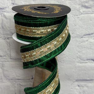 May include: A spool of dark green ribbon featuring a central metallic gold woven band. The ribbon has a smooth, satin-like texture with finished edges and is wrapped around a black circular spool with a gold laurel leaf design printed on top.