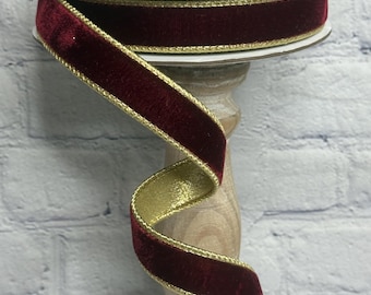 May include: A spool of deep burgundy velvet ribbon featuring metallic gold edges. The ribbon is wound around a black circular spool with white text, displaying a rich, soft texture on the front and a smooth, shimmering gold finish on the back.