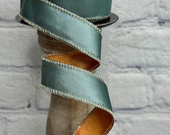 May include: A spool of teal satin ribbon with a gold stitched edge. The ribbon is displayed in a spiral, revealing an orange underside. The ribbon is on a wooden stand against a white brick wall.