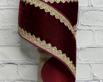 May include: A spool of deep burgundy velvet ribbon featuring a metallic gold scalloped edge trim. The ribbon is wound in a spiral pattern, showcasing the rich red fabric texture contrasted against the intricate, shimmering gold border detailing.
