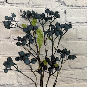 May include: A sprig of artificial blue berries with green leaves and brown stems. The berries are clustered together on the stems.