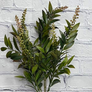 May include: A bouquet of artificial greenery with dark green leaves and small, light green and brown buds. The stems are a light brown color, and the arrangement is in a clear glass vase. The background is a white brick wall.