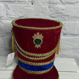 May include: A cylindrical red velvet hat with a flat top and a wide brim. The hat features gold braided trim, a horizontal blue velvet band, and a central gold-toned emblem with a green faux gemstone. Gold tassels hang from the sides.