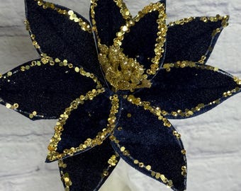 May include: A navy blue fabric flower with gold glitter accents. The flower has multiple petals outlined with gold sequins and a gold glitter center. The flower is displayed in a clear glass vase against a white brick wall.