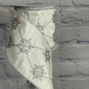 May include: A roll of white ribbon with silver snowflake and leaf embellishments. The ribbon has a silver trim and is displayed against a white brick wall. The ribbon is ideal for holiday decorations and gift wrapping.