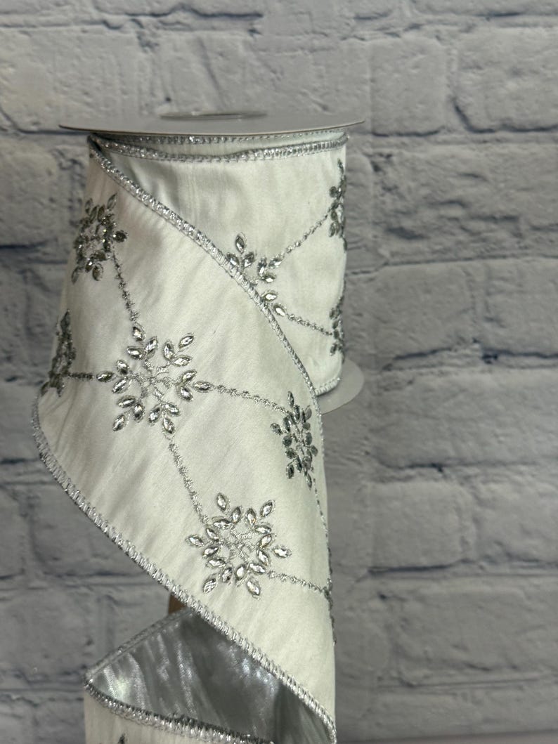 May include: A roll of white ribbon with silver snowflake and leaf embellishments. The ribbon has a silver trim and is displayed against a white brick wall. The ribbon is ideal for holiday decorations and gift wrapping.