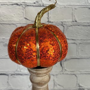 Puede incluir: Una calabaza naranja con lentejuelas, detalles dorados y un tallo dorado brillante. La calabaza decorativa está adornada con cinta dorada y se apoya sobre un candelabro de madera. Una decoración festiva de otoño.
