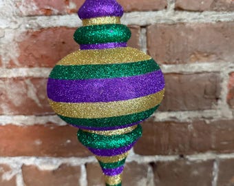 May include: A purple, green, and gold glitter Christmas ornament with a pointed tip. The ornament is striped with gold, purple, and green glitter.