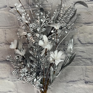 K&ouml;nnte beinhalten: Ein Strau&szlig; k&uuml;nstlicher Blumen in einer durchsichtigen Glasvase. Das Arrangement besteht aus silberfarbenen Bl&auml;ttern, Beeren und Stielen, mit wei&szlig;en Bl&uuml;ten. Der Hintergrund ist eine wei&szlig;e Backsteinmauer.