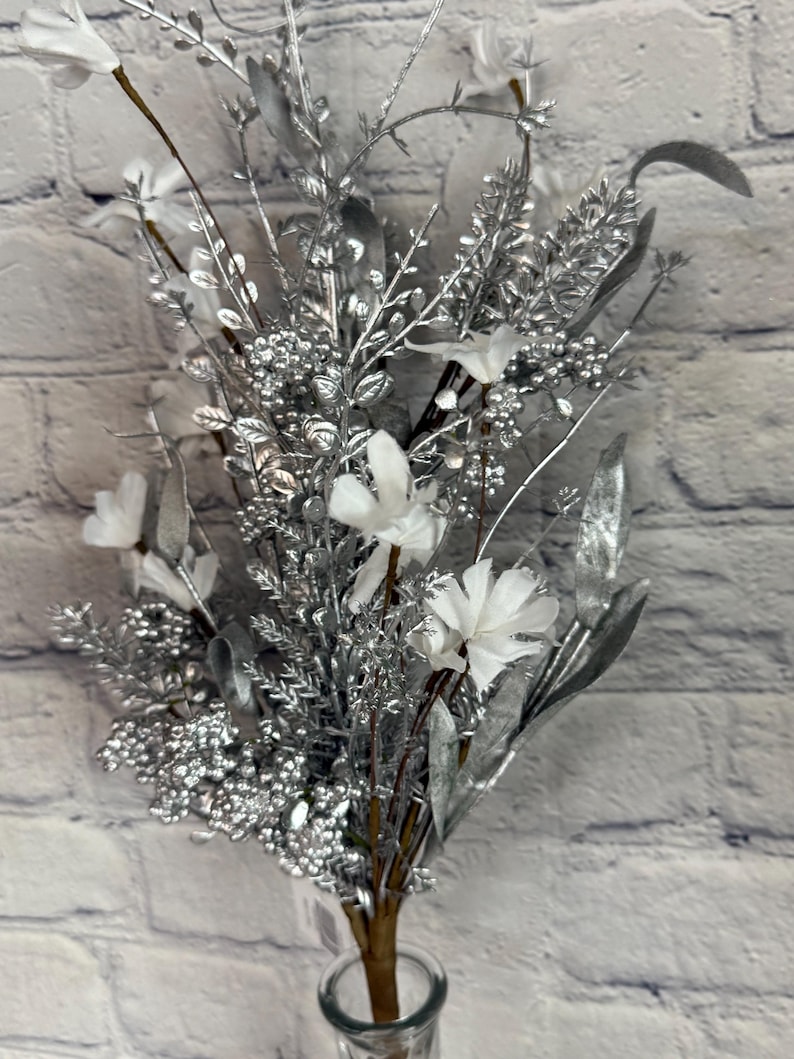 K&ouml;nnte beinhalten: Ein Strau&szlig; k&uuml;nstlicher Blumen in einer durchsichtigen Glasvase. Das Arrangement besteht aus silberfarbenen Bl&auml;ttern, Beeren und Stielen, mit wei&szlig;en Bl&uuml;ten. Der Hintergrund ist eine wei&szlig;e Backsteinmauer.