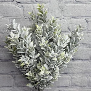 May include: A bundle of artificial greenery with small, light green leaves dusted with white, resembling frost. The leaves are clustered together, creating a full, textured appearance. The stems are bound together, and the background is a gray brick wall.