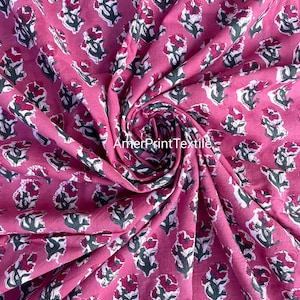 May include: Pink fabric with a repeating floral pattern. The flowers are a deep red with white and green accents. The fabric is folded, showing the texture and pattern. The text "AmerPrintTextile" is visible in the bottom left corner.