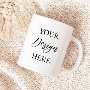 May include: White ceramic mug with the text "YOUR Design HERE" in black script and block letters. The mug is on a cream-coloured knitted surface, with a cream-coloured macrame detail in the upper left corner. The mug has a curved handle.