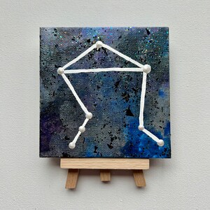 May include: A small canvas painting of the Libra constellation on a wooden easel. The constellation is painted in white with white dots on a dark blue and purple background with glitter.