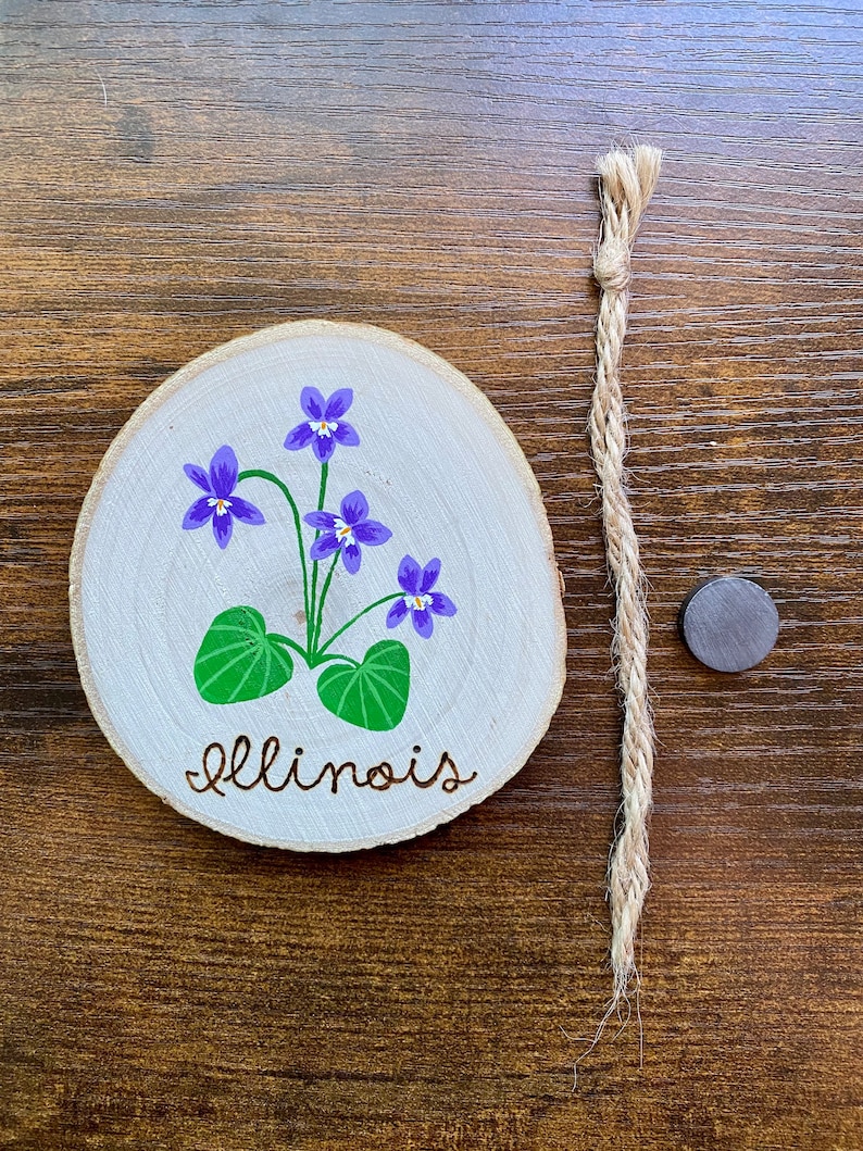 May include: A wooden round slice with a painted purple flower and green leaves. The word "Illinois" is written in cursive on the wood.