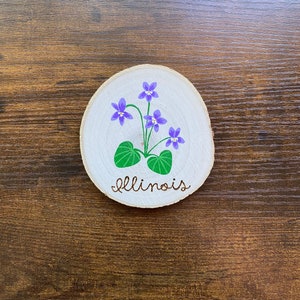 May include: A round wooden slice with a painted design of purple flowers and green leaves. The word "Illinois" is written in cursive below the flowers.