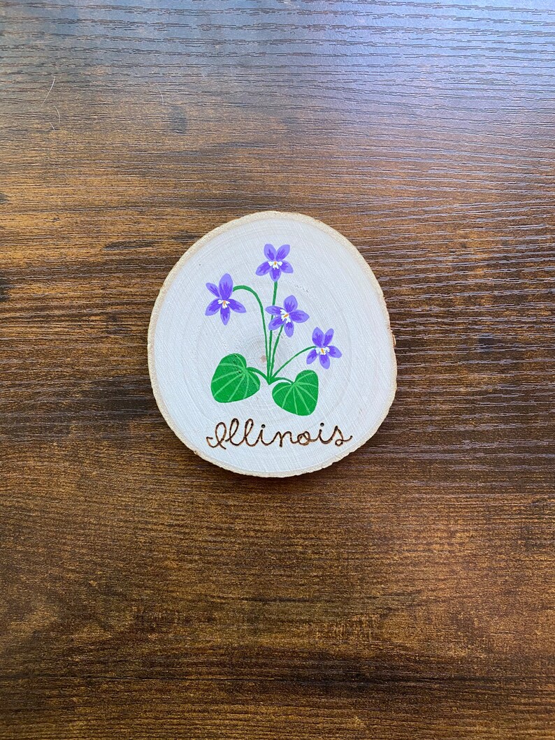 May include: A round wooden slice with a painted design of purple flowers and green leaves. The word "Illinois" is written in cursive below the flowers.