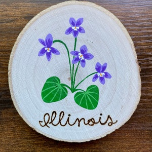 May include: A round wooden slice with a painted image of purple and white violets with green leaves and the word "Illinois" written in brown paint.