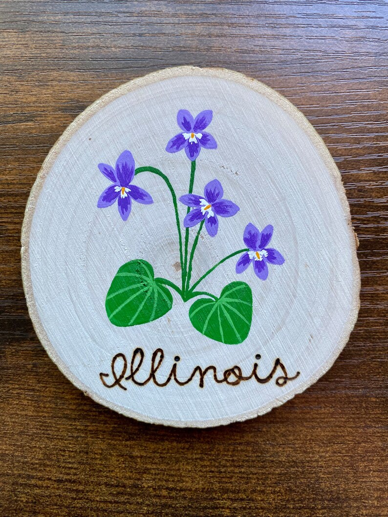 May include: A round wooden slice with a painted image of purple and white violets with green leaves and the word "Illinois" written in brown paint.