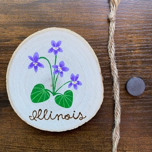 May include: A wooden round slice with a painted purple violet flower and green leaves. The words "Illinois" are written in cursive below the flower. The slice is attached to a piece of twine.