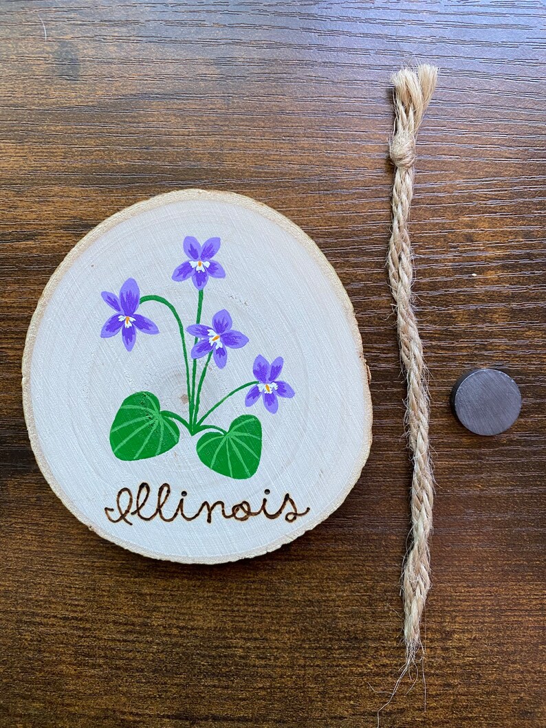 May include: A wooden round slice with a painted purple violet flower and green leaves. The words "Illinois" are written in cursive below the flower. The slice is attached to a piece of twine.