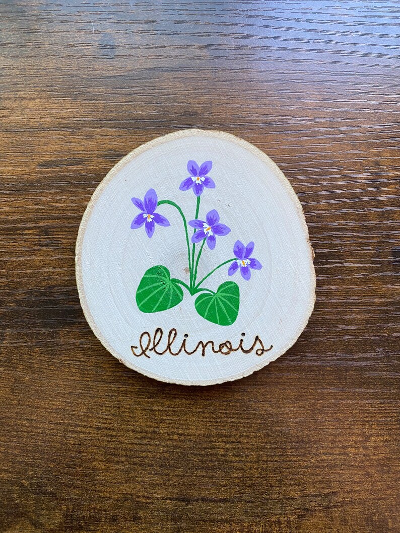 May include: A round wooden slice with a painted purple violet flower with green leaves and the word "Illinois" written in cursive.