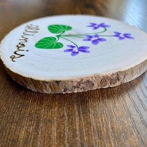 May include: A round wooden slice with the word "Illinois" burnt into the wood. The slice is painted with purple flowers and green leaves.