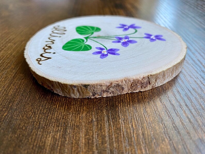 May include: A round wooden slice with the word "Illinois" burnt into the wood. The slice is painted with purple flowers and green leaves.