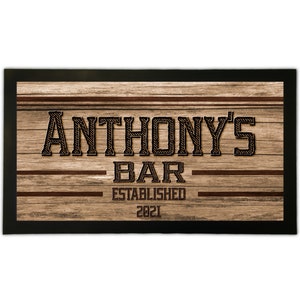 Puede incluir: Un letrero de madera con el texto "Anthony's Bar Establecido 2021" en una fuente de cuerda negra y marrón. El letrero tiene un marco negro y un fondo de veta de madera.