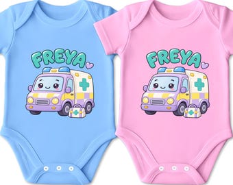 Könnte beinhalten: Zwei kurzärmelige Babybodys, einer in Hellblau und einer in Hellrosa, jeweils mit einer Cartoon-Krankenwagen-Grafik mit lächelndem Gesicht, einem kleinen Erste-Hilfe-Kasten und dem Namen FREYA in großen türkisfarbenen Buchstaben darüber.