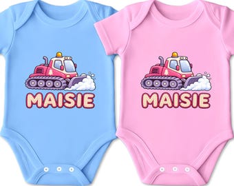 Könnte beinhalten: Zwei kurzärmelige Babybodys, einer in Hellblau und einer in Hellrosa, jeweils mit einer Grafik eines roten Pistenfahrzeugs, das Schnee schiebt, und dem Namen MAISIE, der in großen, fetten, hellen Buchstaben mit dunkler Umrandung darunter steht.