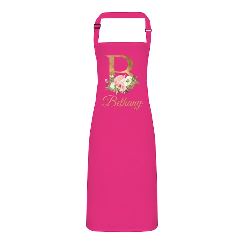 Op de afbeelding: Roze keukenschort met een gouden bloemmotief initial "B" en de naam "Bethany" bedrukt op de voorkant.
