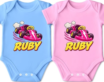 Könnte beinhalten: Zwei kurzärmelige Babybodys, einer in Hellblau und einer in Hellrosa, jeweils mit einer Grafik eines pinkfarbenen Karts mit weißem Blitz, weißen Abgaswolken und dem Namen RUBY in fetten gelben Buchstaben unter dem Fahrzeug.