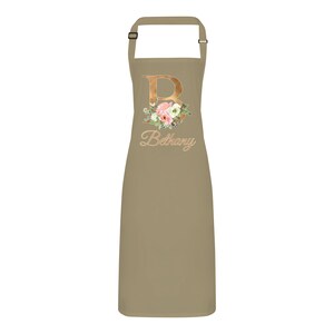 Op de afbeelding: Een beige schort met een gouden bloemenontwerp met de letter "B" en de naam "Bethany" in goud.
