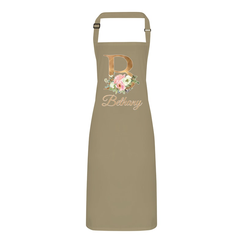Op de afbeelding: Een beige schort met een gouden bloemenontwerp met de letter "B" en de naam "Bethany" in goud.