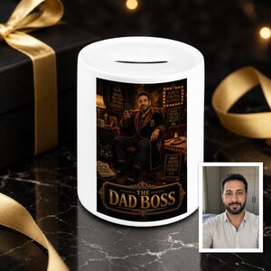 Könnte beinhalten: Eine weiße Keramik-Spardose mit einer grafischen Illustration einer Person im Morgenmantel, die in einem Sessel sitzt, mit dem Text THE DAD BOSS. Das Design enthält kleine Schilder mit Text und ein kleines eingefügtes Foto einer bärtigen Person.