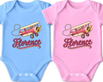 Könnte beinhalten: Zwei kurzärmelige Babybodys, einer in Hellblau und einer in Hellrosa, jeweils mit einer Grafik eines roten Doppeldeckers mit Propeller und einer gepunkteten Flugbahnlinie sowie dem Namen Florence in einer roten Schreibschrift.