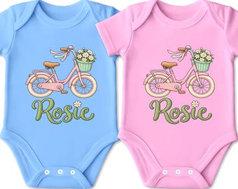 Könnte beinhalten: Zwei kurzärmelige Babybodys, einer in Hellblau und einer in Hellrosa, jeweils mit einer Grafik eines rosa Fahrrads mit einem Korb voller weißer Gänseblümchen am Lenker und dem Namen Rosie in grüner Schreibschrift unter dem Fahrrad.