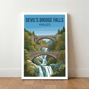 May include: A framed print of Devil's Bridge Falls in Wales. The image shows three stone bridges spanning a waterfall, set amidst green trees and foliage. The sky is blue, and the text "DEVIL'S BRIDGE FALLS WALES" is displayed at the top of the artwork.