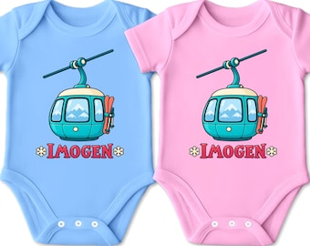 Könnte beinhalten: Zwei kurzärmelige Babybodys, einer in Hellblau und einer in Hellrosa, jeweils mit einer Grafik einer türkisfarbenen Skigondel mit roten Skiern an der Seite und dem Namen IMOGEN in roten Buchstaben zwischen zwei weißen Schneeflocken-Symbolen.