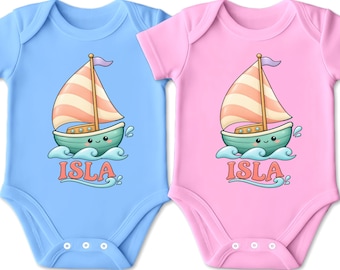 Könnte beinhalten: Zwei kurzärmelige Babybodys, einer in Hellblau und einer in Hellrosa, jeweils mit einer Grafik eines lächelnden Segelboots auf blauen Wellen und dem Namen ISLA in einer orangen Retro-Schrift unter dem Boot, inklusive Druckknöpfen aus Metall.