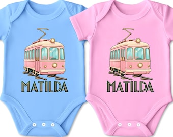 Könnte beinhalten: Zwei kurzärmelige Babybodys, einer in Hellblau und einer in Hellrosa, jeweils mit einer grafischen Abbildung einer rosa Vintage-Straßenbahn auf Schienen und dem Namen MATILDA in dunkelgrünen Großbuchstaben unter der Straßenbahn.