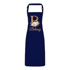 Op de afbeelding: Donkerblauwe schort met een gouden letter "B" en een bloemenontwerp. De naam "Bethany" staat in gouden letters onder de letter.