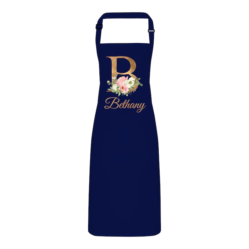 Op de afbeelding: Donkerblauwe schort met een gouden letter "B" en een bloemenontwerp. De naam "Bethany" staat in gouden letters onder de letter.