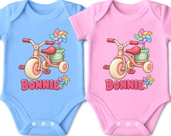 Könnte beinhalten: Zwei kurzärmelige Bodys, einer hellblau und einer hellrosa, jeweils mit einer Grafik eines rosafarbenen Dreirads mit einem grünen Eimer, der ein buntes Windrad enthält. Der Name BONNIE ist in rosa Blasenbuchstaben unter dem Dreirad gedruckt.